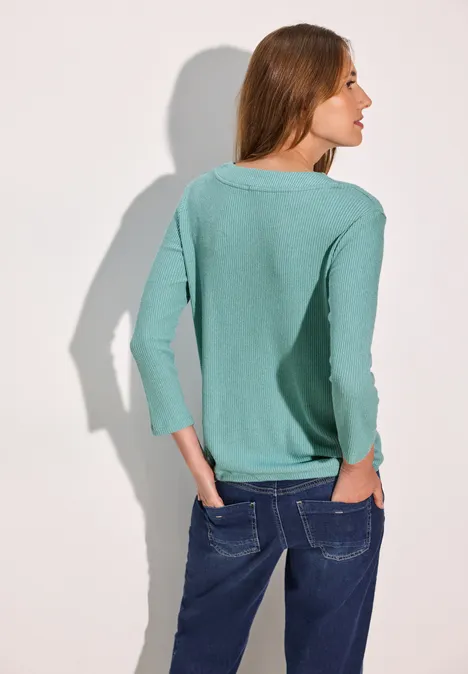 Frau mit hellgrünem Pullover und Jeans, steht rückwärts vor hellem Hintergrund.